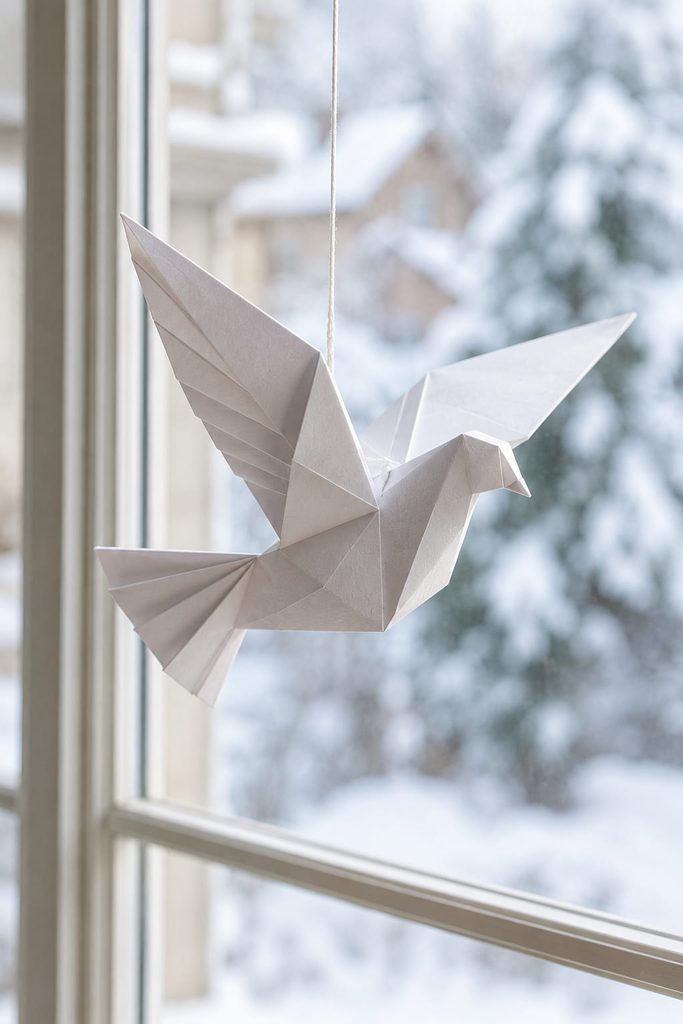 White folded-paper dove ornament hanging level in front of a winter window.