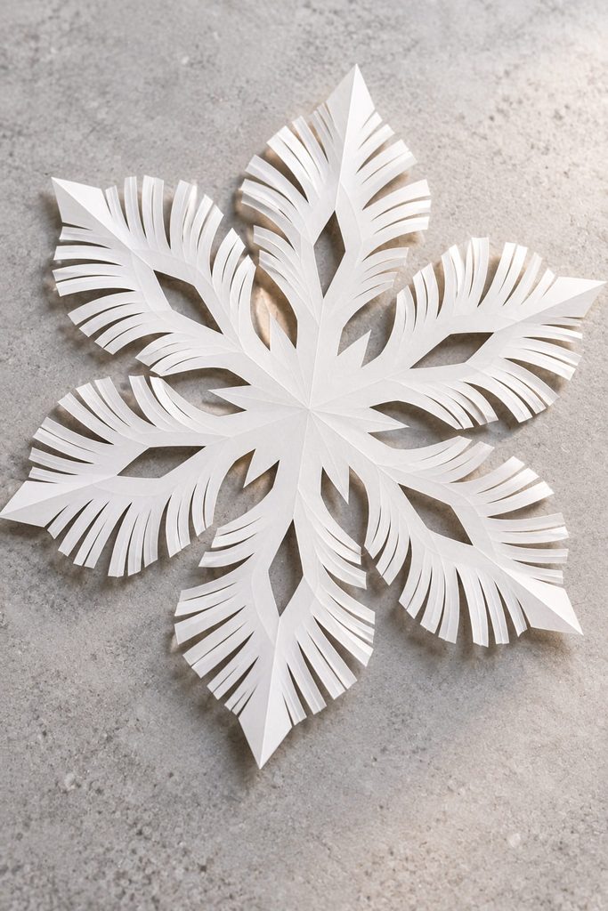 Feathered paper snowflake with narrow fringe slits and a solid center on a pale gray surface.